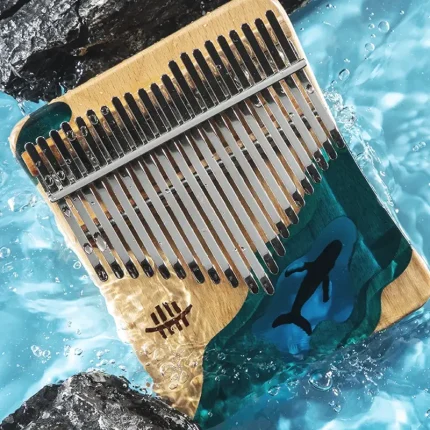 A dynamic shot of the ergonomic Hluru 21 key kalimba with the whale design, with water splashing around it between dark rocks.