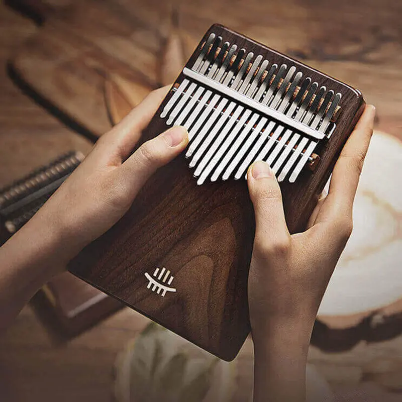 A close-up shot of two hands holding and about to play the Hluru 17 key sloped solid body kalimba, showcasing its tiger striped walnut wood.