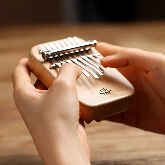 A pair of hands holding and playing the compact Hluru Mini 8 Key Kalimba, crafted from light-colored maple wood, demonstrating its comfortable size.