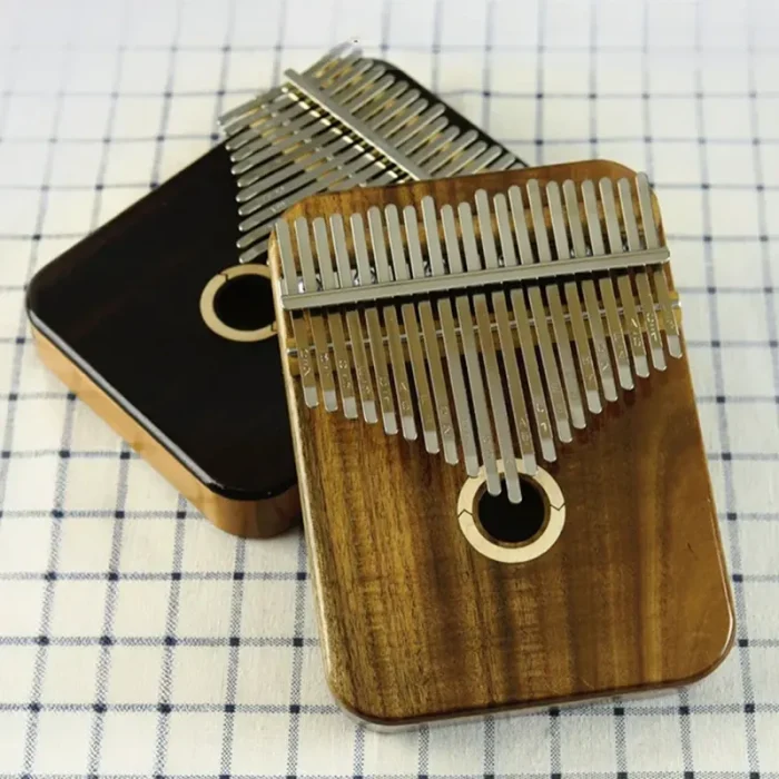 Two LOOP kalimbas, a 17/21 key model in bright Acacia wood and another in dark Rosewood, shown together on a checkered cloth. Caption: Choose your voice: the bright, clear tone of Acacia (front) or the deep, warm resonance of Rosewood (back).