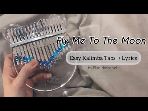 A hand plays a clear acrylic kalimba labeled with blue and white notes. Text reads "fly-me-to-the-moon-kalimba-tabs-cover-2: Easy Kalimba Tabs + Lyrics by Elisa Nathaniel" over a soft, neutral background.