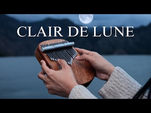 Hands playing a kalimba at dusk by a lake and mountains with a full moon, featuring large "CLAIR DE LUNE" text above. Product: clair-de-lune-kalimba-tabs-cover.